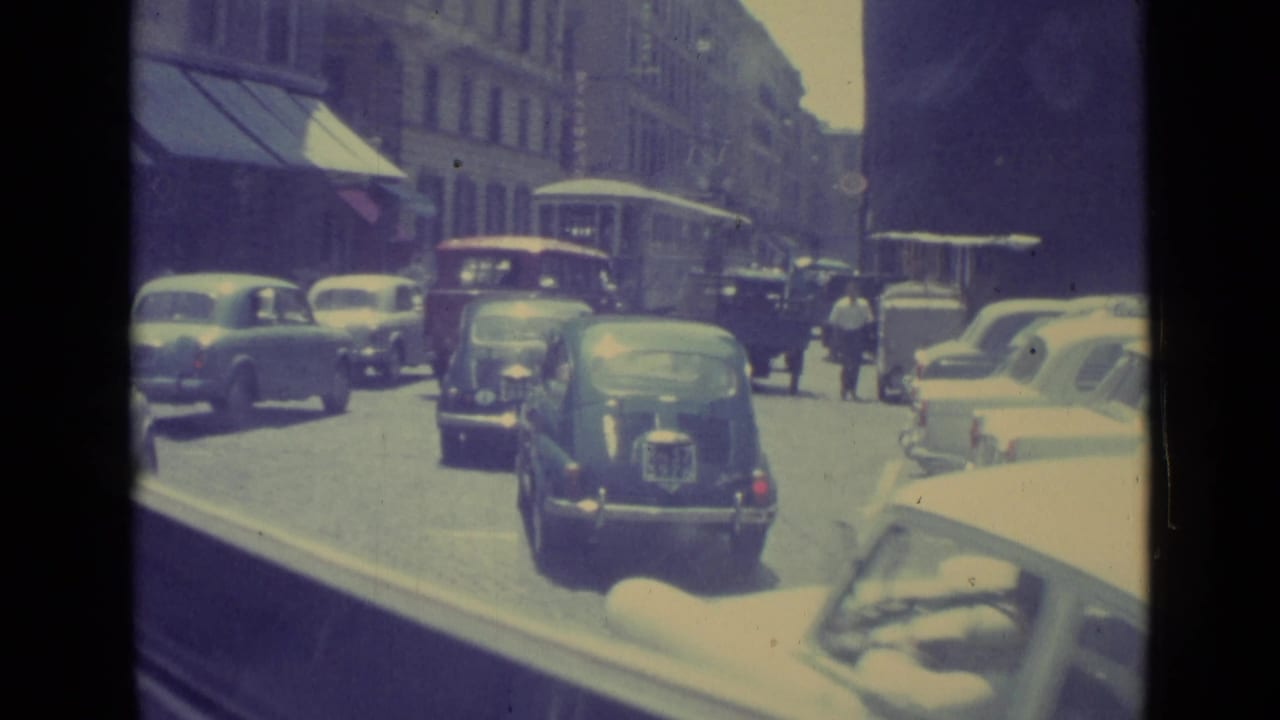 Busy city street with vintage cars, pedestrians, awnings, and a distant bus or trolley.