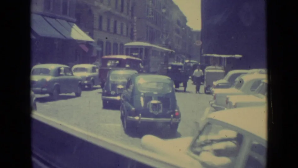 1962 Vatican City street view with period cars moving past pedestrians, storefront awnings, and tall buildings.