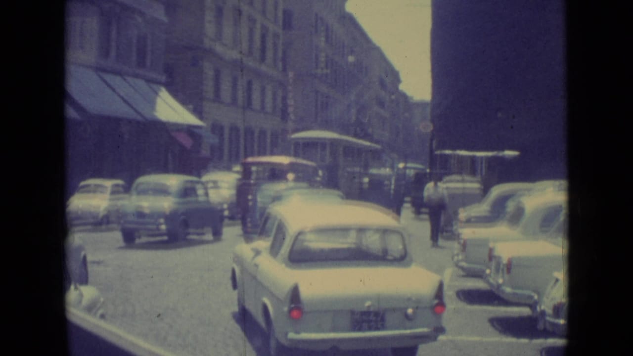 A light-colored sedan moves through a 1962 Vatican City street scene with pedestrians and period traffic.