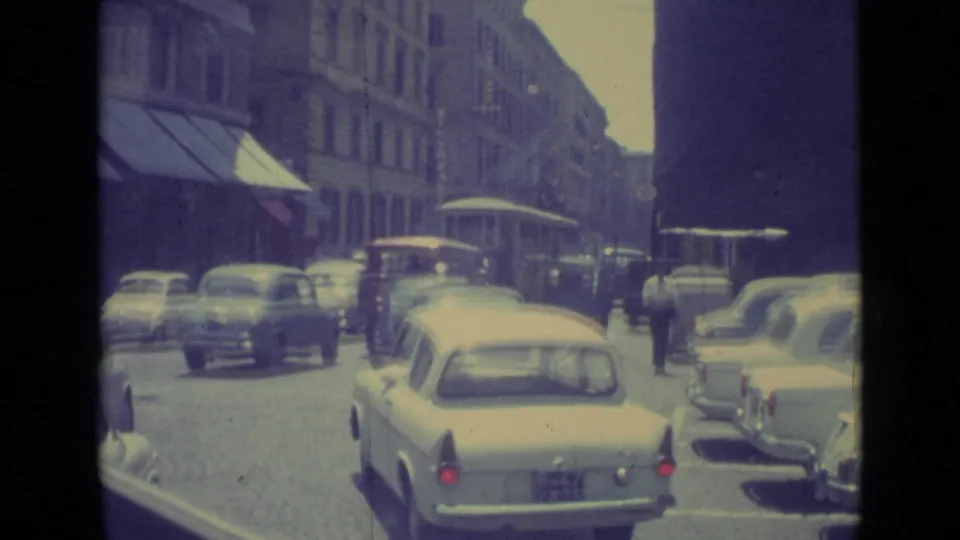 A light-colored sedan moves through a 1962 Vatican City street scene with pedestrians and period traffic.