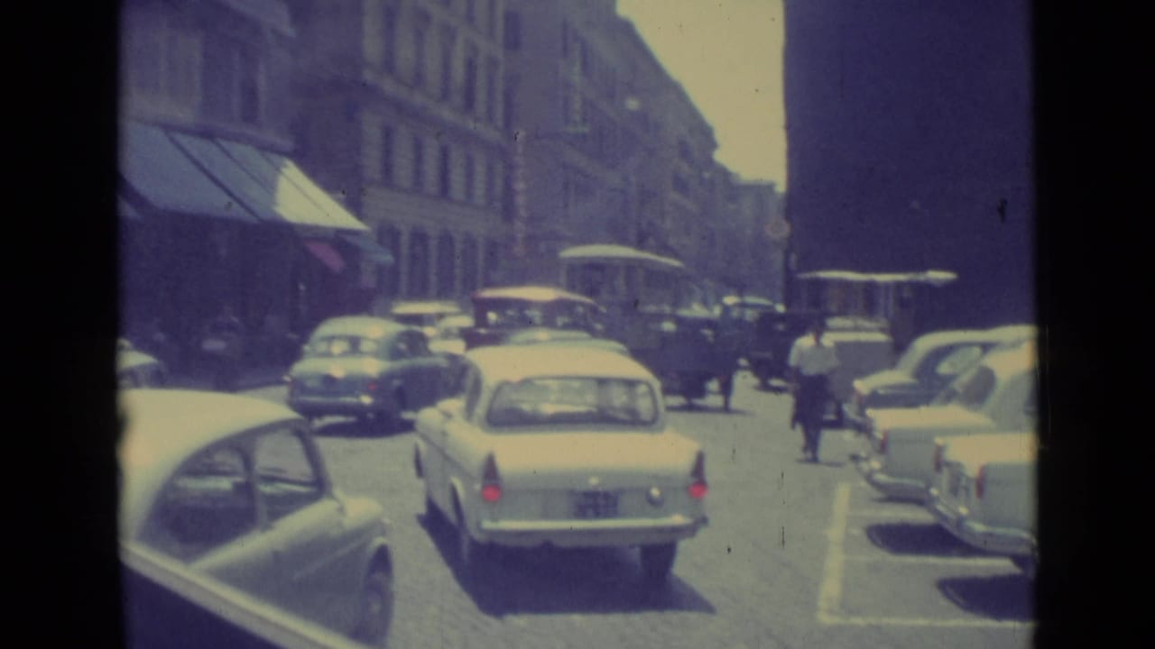 Rear view of a light sedan amid 1962 Vatican City traffic, with pedestrians and parked cars along the street.