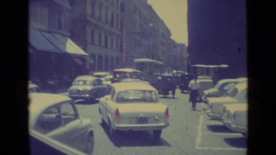 Rear view of a light sedan amid 1962 Vatican City traffic, with pedestrians and parked cars along the street.
