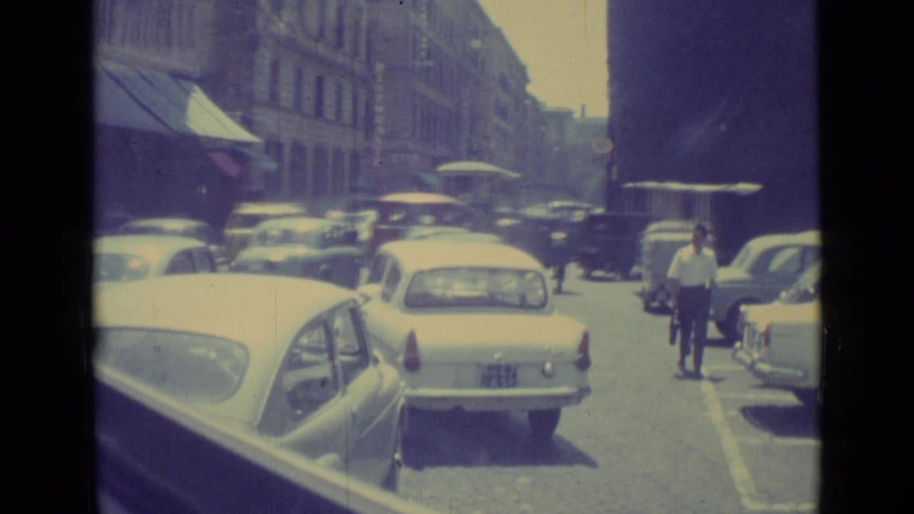 1962 Vatican City traffic scene with a light sedan, parked cars, pedestrians, awnings, and a cyclist near the curb.