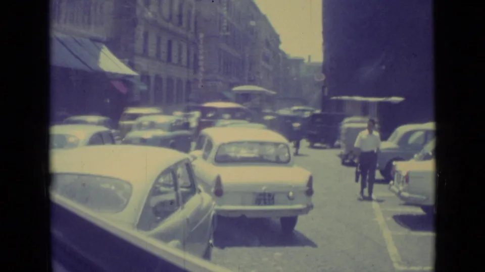 1962 Vatican City traffic scene with a light sedan, parked cars, pedestrians, awnings, and a cyclist near the curb.