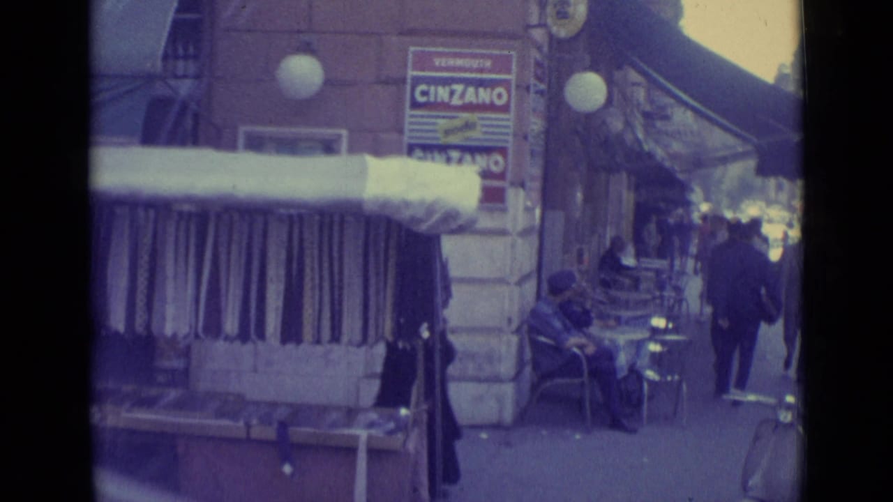 1962 Vatican City sidewalk scene showing outdoor seating, a seated person, passing pedestrians, a shop awning, and Cinzano signage.