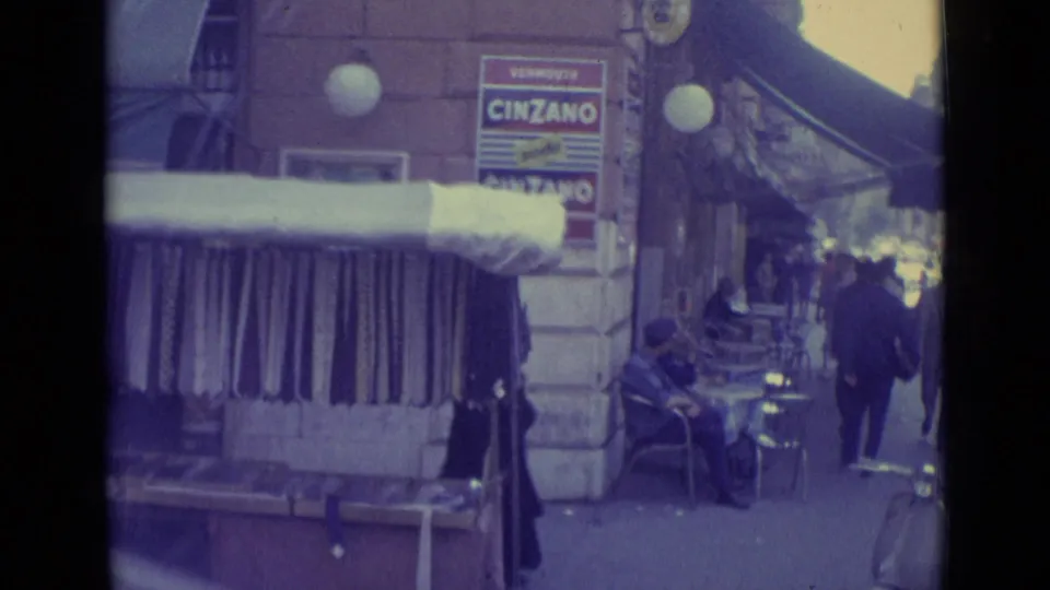 1962 Vatican City sidewalk scene showing outdoor seating, a seated person, passing pedestrians, a shop awning, and Cinzano signage.