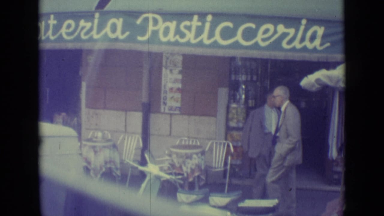 1962 Vatican City vehicle POV showing a man walking past cafe seating beneath a gelateria and pasticceria awning.