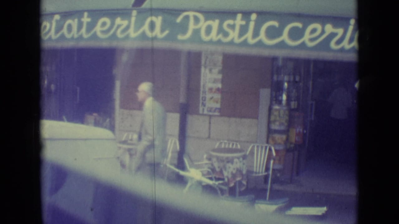 1962 Vatican City street frontage with a pedestrian, outdoor cafe chairs, shop doorway, and pasticceria awning seen from a vehicle.