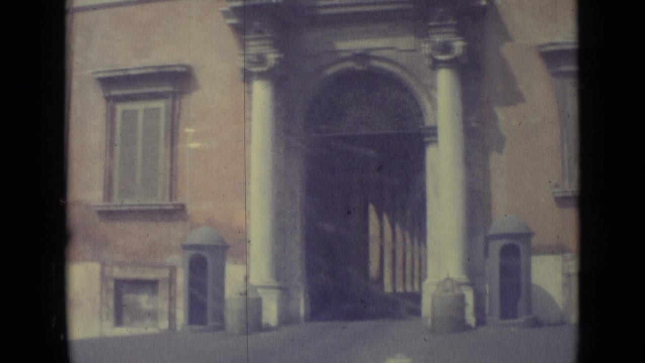 1962 Vatican City frame with a formal arched doorway, flanking columns, shuttered windows, and warm light across the stone exterior.