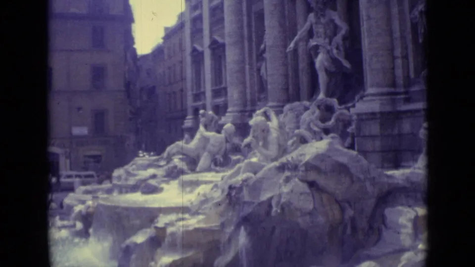 1962 8mm view of Trevi Fountain stone sculptures, columns, and cascading water with surrounding buildings in the background.