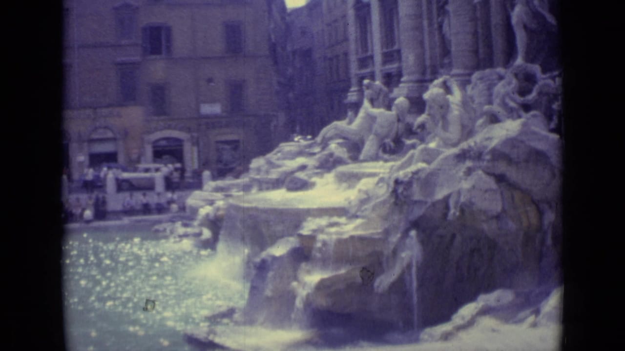 1962 home-movie frame showing tourists around the Trevi Fountain basin, with carved rocks and flowing water in view.