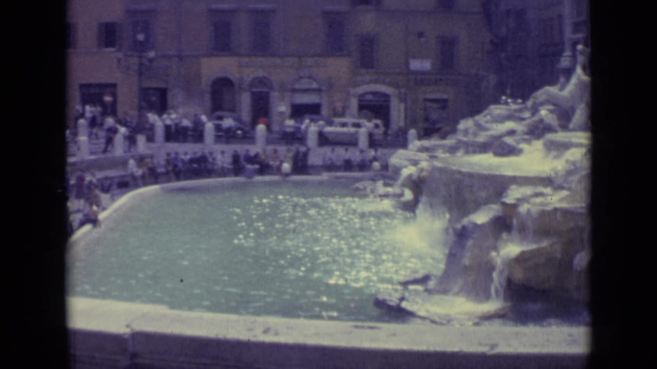 Cataloged 1962 8mm view of the Trevi Fountain basin, surrounding tourists, stone cascades, and plaza buildings.