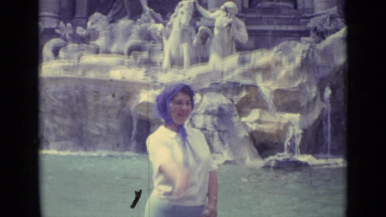 1962 8mm travel frame showing a young girl in a light top and headscarf making a coin-toss gesture near the Trevi Fountain pool.