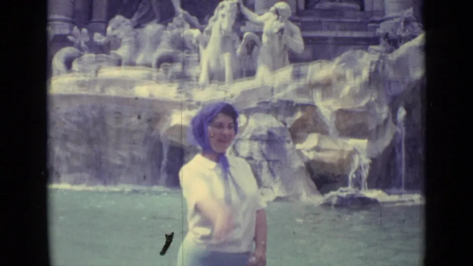 1962 8mm travel frame showing a young girl in a light top and headscarf making a coin-toss gesture near the Trevi Fountain pool.