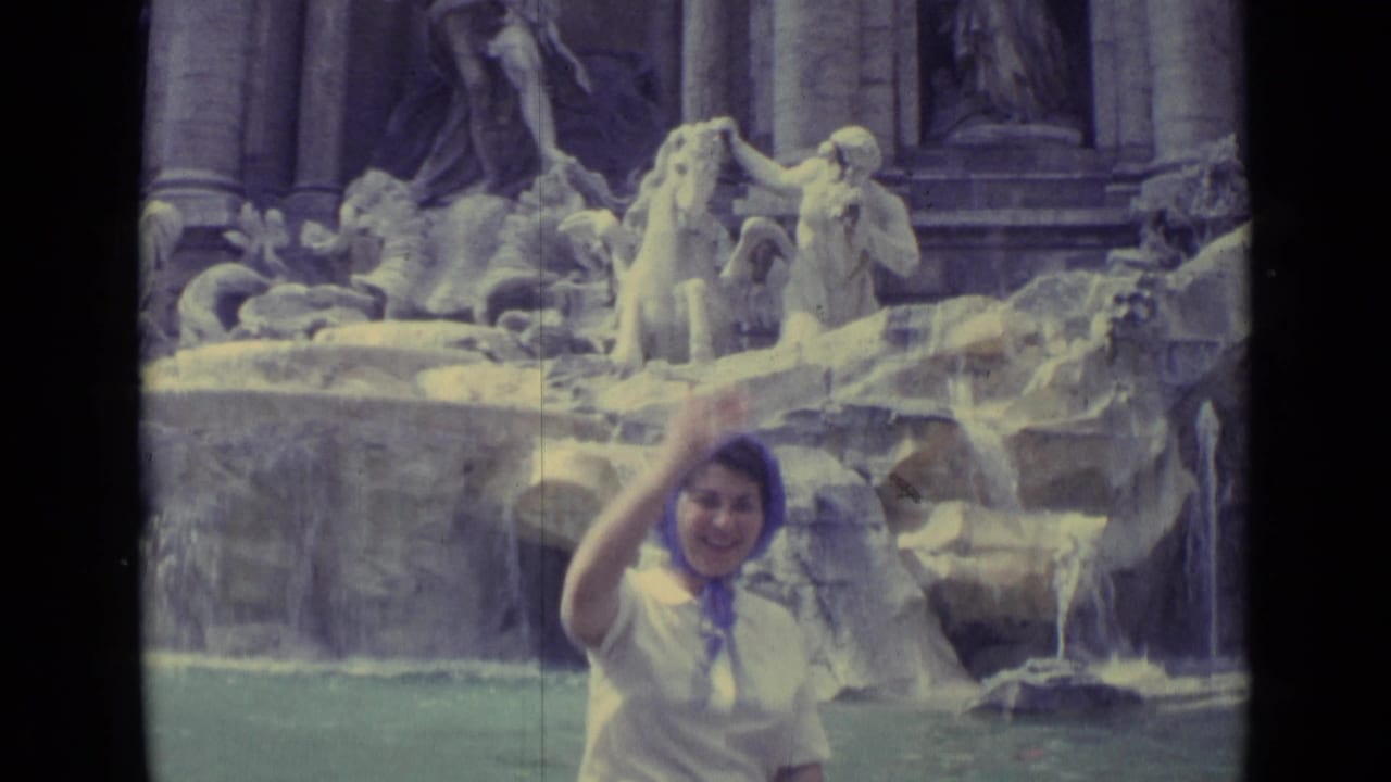 Woman in a purple headscarf waves in front of a large stone fountain.