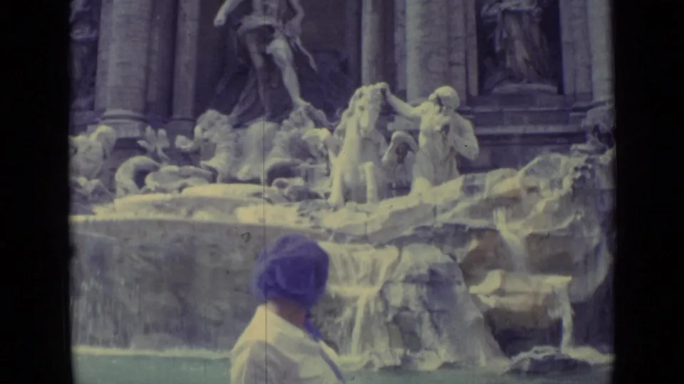 The woman faces the fountain, her purple headscarf visible against the stone sculptures and cascading water.
