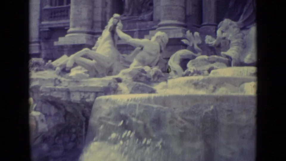Aged 8mm frame showing a monumental fountain facade with sculpted figures, horses, columns, and water spilling forward.