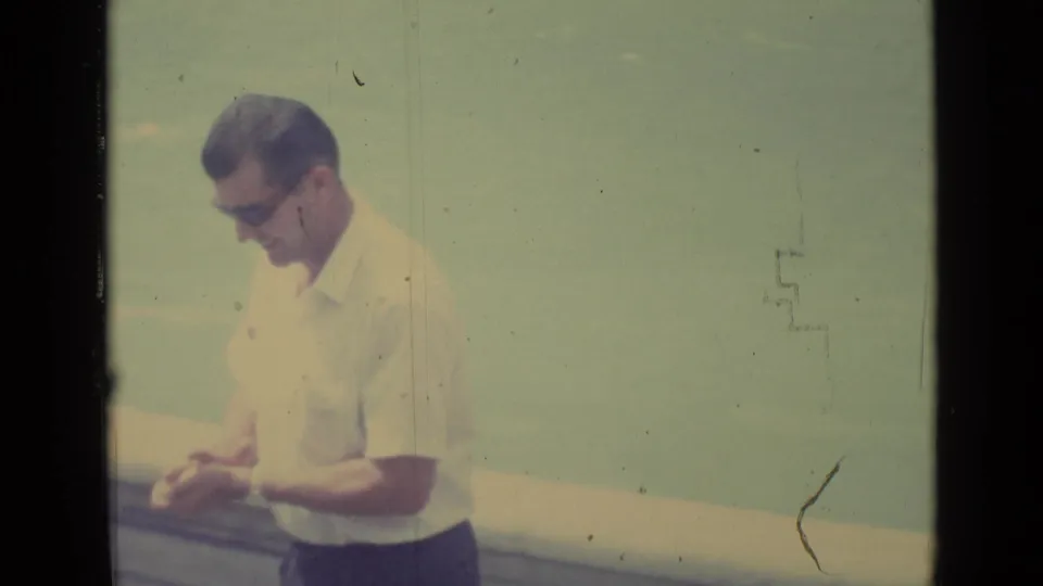 The same adult man in 1962 Vatican City footage looks down at his hands, with a pale open background behind him.