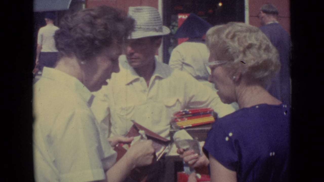 1962 Vatican City home-movie frame of a straw-hatted street vendor offering small items to two women near storefront windows.