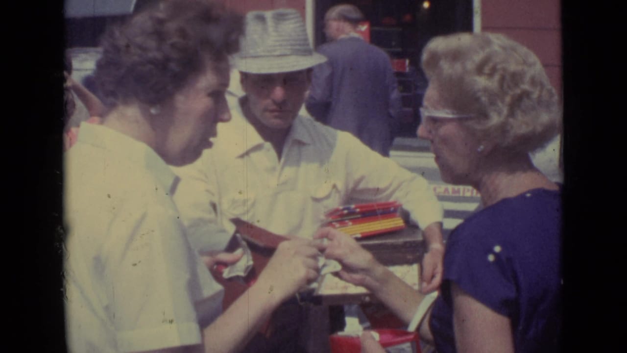 In the Vatican City street scene, two women examine a red case or wallet beside a vendor carrying a tray of small goods.