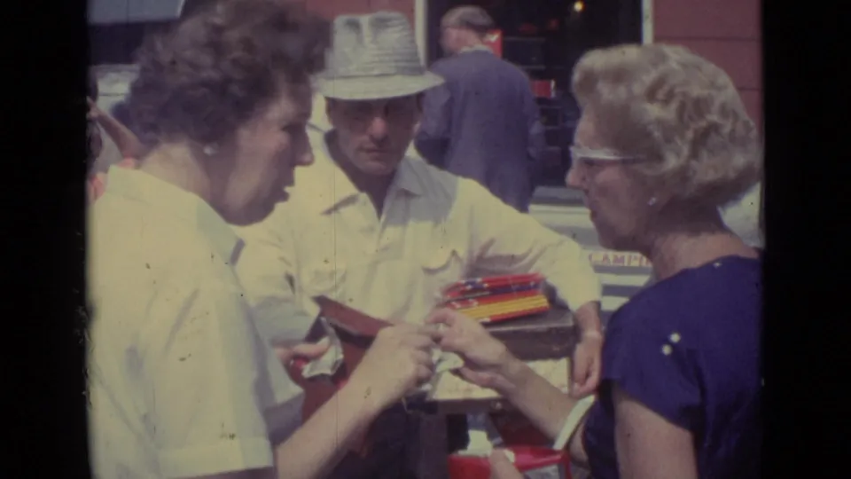 In the Vatican City street scene, two women examine a red case or wallet beside a vendor carrying a tray of small goods.