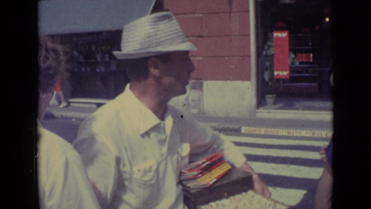 Street vendor in a straw hat stands by a crosswalk holding goods as pedestrians pass.