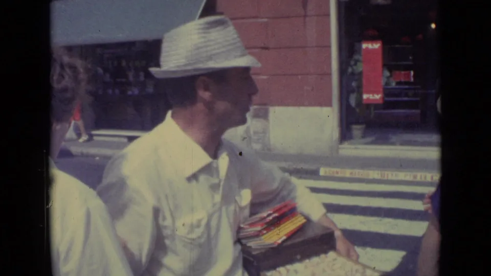 In 1962 Vatican City footage, a straw-hatted vendor stands beside a crosswalk with small goods while pedestrians move near a red storefront.