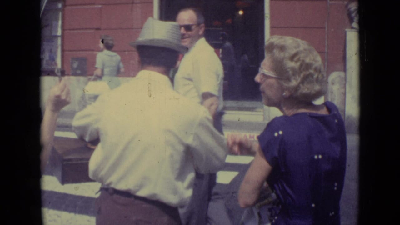From behind the vendor, pedestrians cluster near a crosswalk while a woman in a purple dress watches the street exchange.