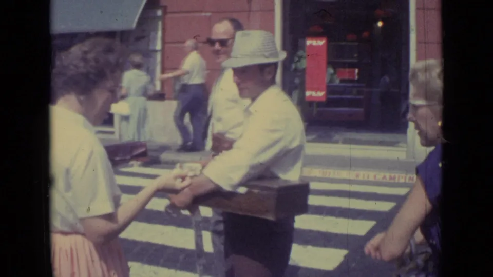 A woman reaches toward the vendor's hand or goods as he carries a wooden box on a Vatican City street in 1962.