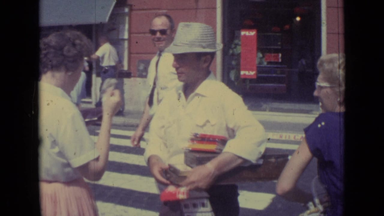 1962 travel footage frame showing a hat-wearing man with boxed items near pedestrians and storefronts in the Vatican City clip.