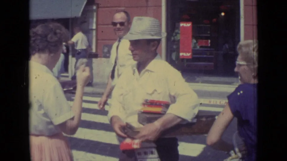 1962 travel footage frame showing a hat-wearing man with boxed items near pedestrians and storefronts in the Vatican City clip.