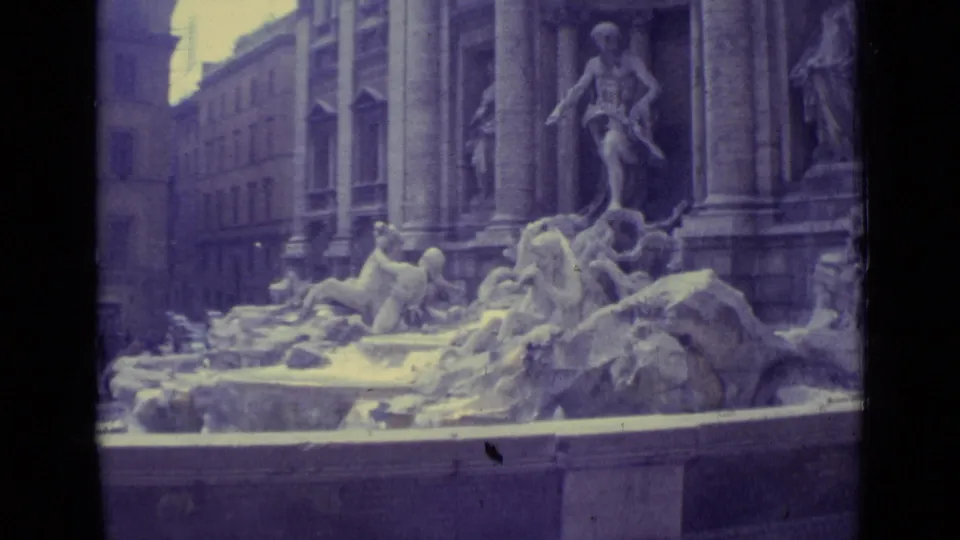 Roman landmark view from the clip showing the Trevi Fountain sculptures, columns, and nearby buildings in handheld 8mm footage.