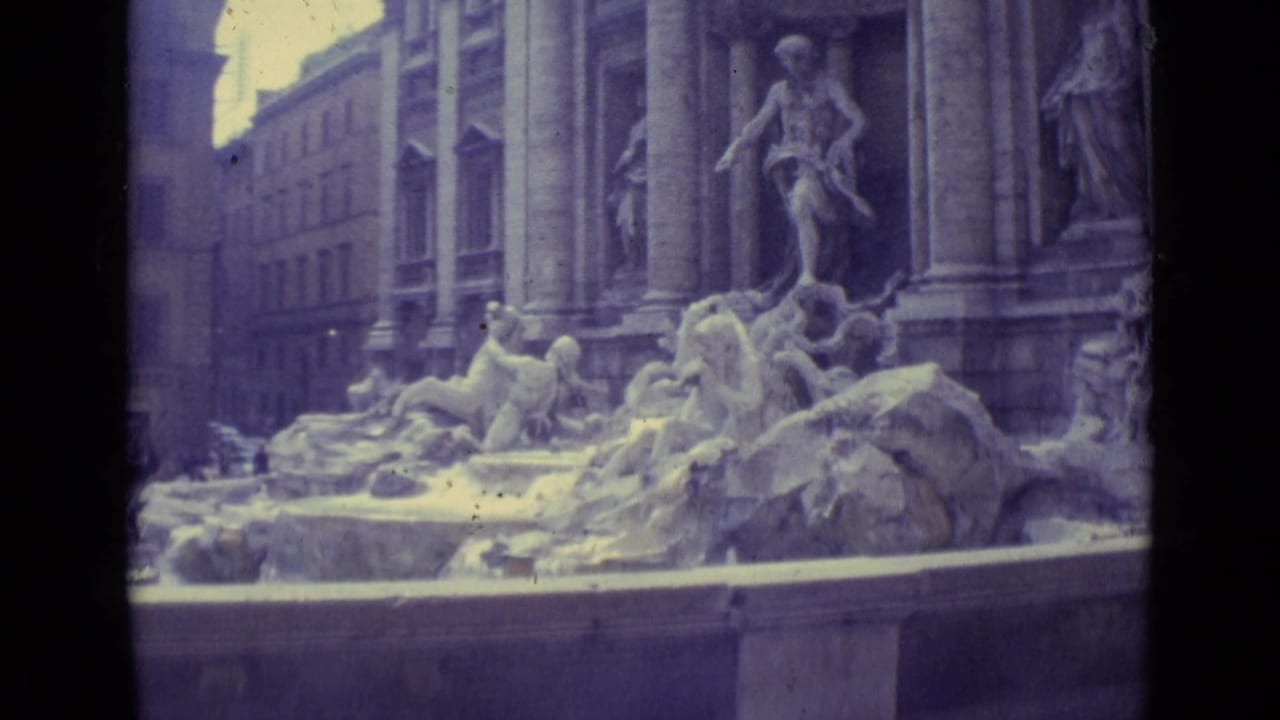 Handheld 8mm view of the Trevi Fountain in Rome, showing sculpted figures, the basin edge, columns, and adjacent buildings.