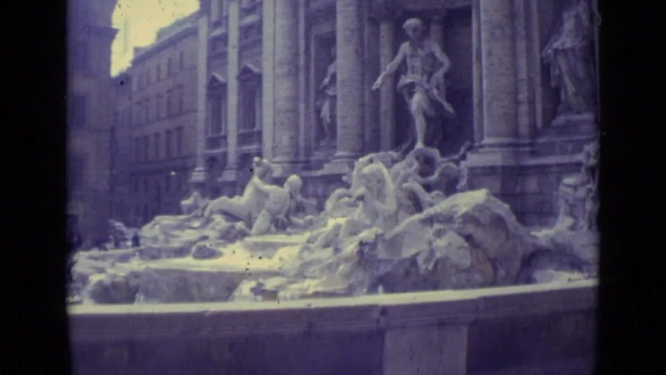 Handheld 8mm view of the Trevi Fountain in Rome, showing sculpted figures, the basin edge, columns, and adjacent buildings.