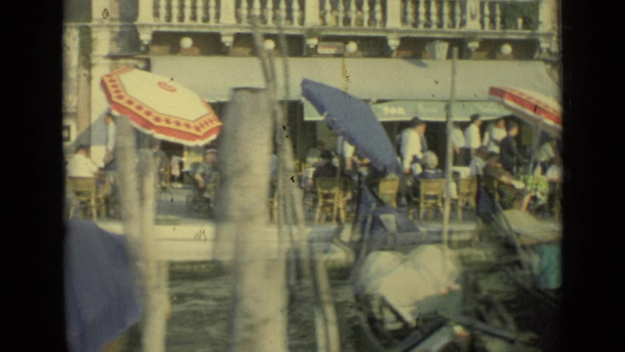 Cafe umbrellas and diners beside a Venice canal with gondolas in the foreground.
