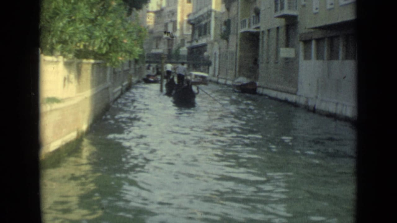 1962 home-movie view down a narrow Venice canal, with gondolas, rippled water, and shaded buildings on both sides.