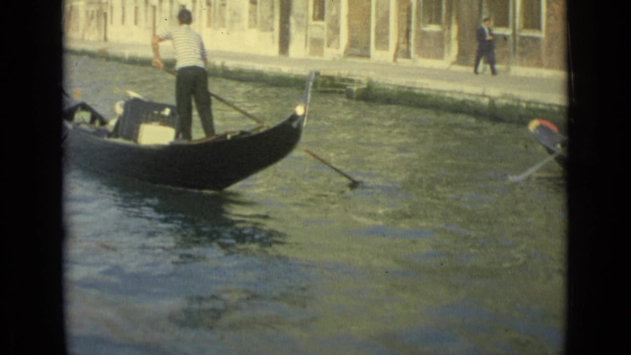 1962 Venice home-movie frame showing a striped-shirt gondolier rowing passengers and luggage past canal-side buildings.