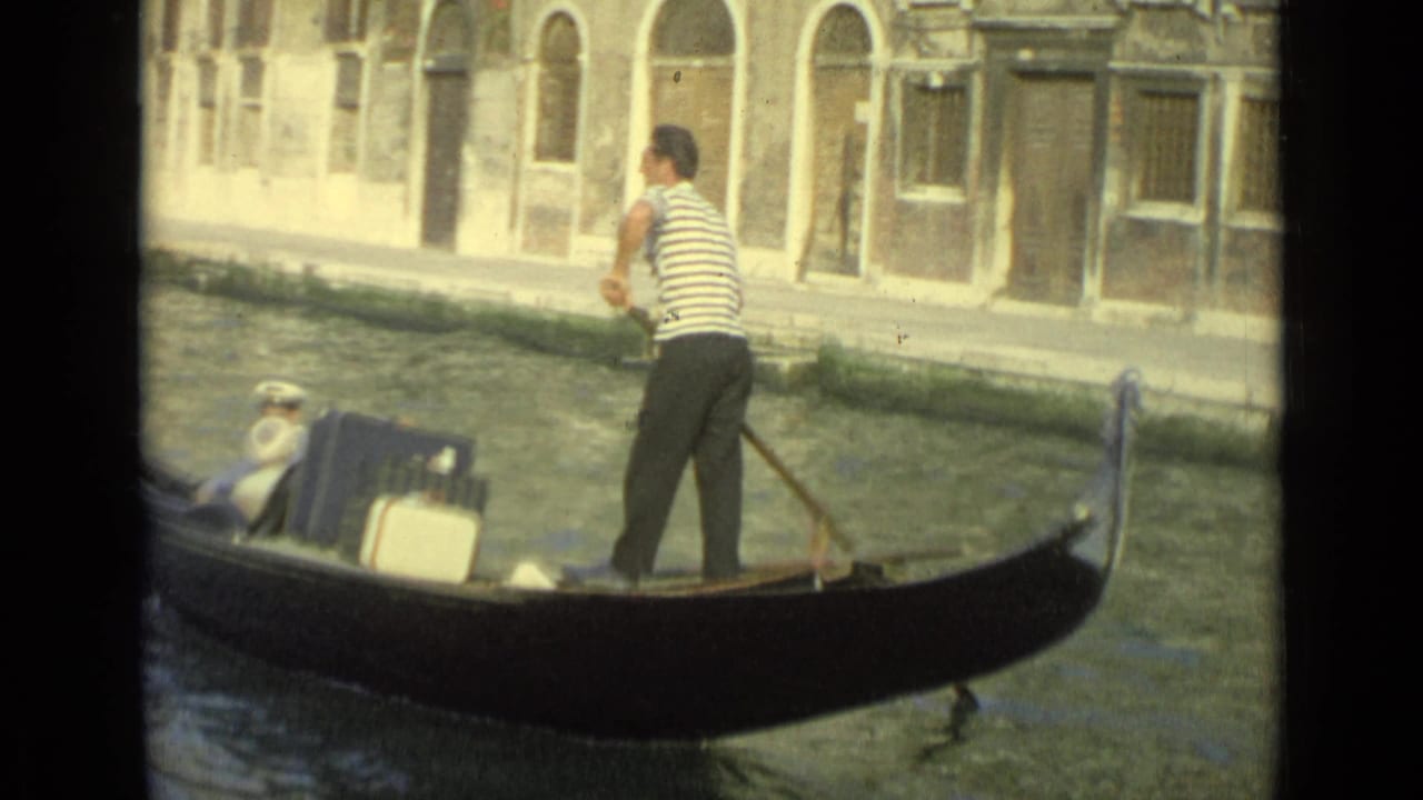Closer 1962 Venice frame showing a striped gondolier, passengers, a blue suitcase, and canal-side stonework.