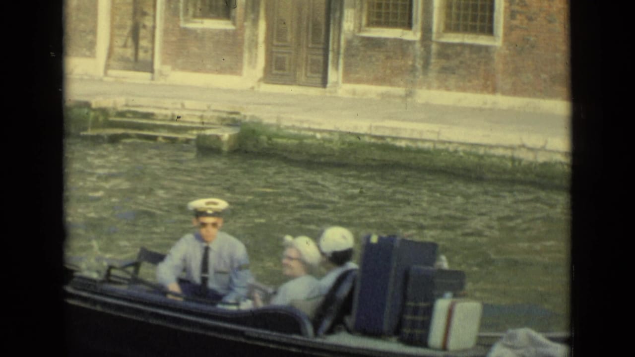 Passengers, including a man in a captain-style hat, sit in a Venice gondola with luggage beside the canal wall.