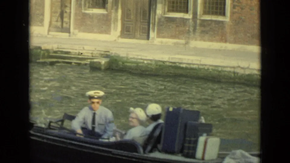 Passengers, including a man in a captain-style hat, sit in a Venice gondola with luggage beside the canal wall.