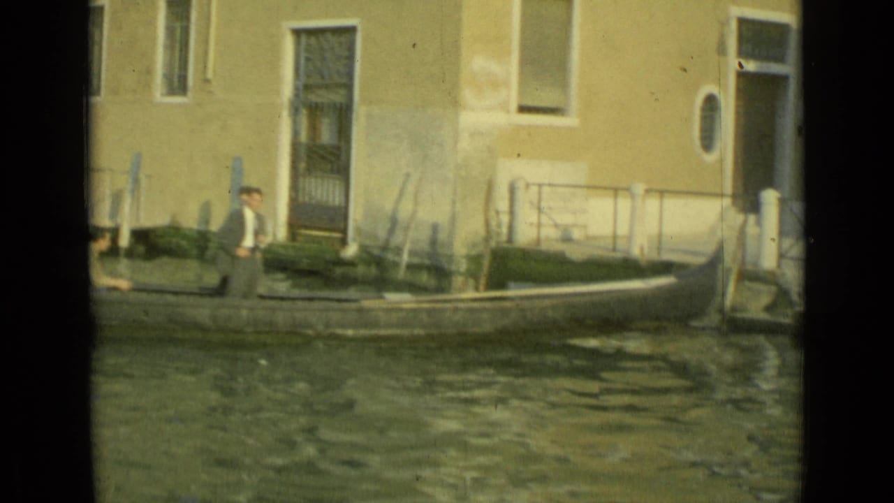 A small boat moves through a Venice canal while a man waits near mooring posts and a worn building facade.