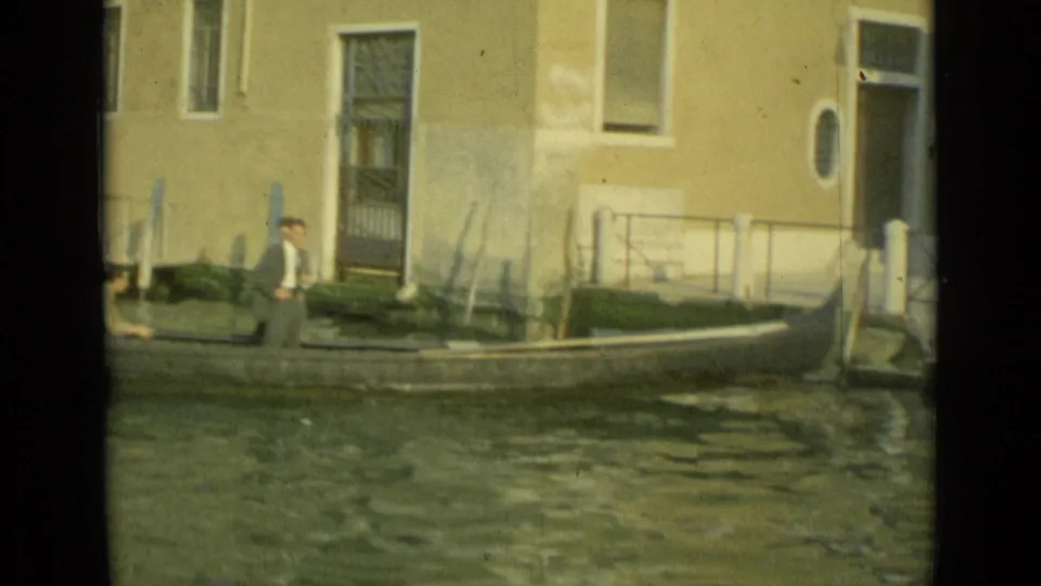 A small boat moves through a Venice canal while a man waits near mooring posts and a worn building facade.