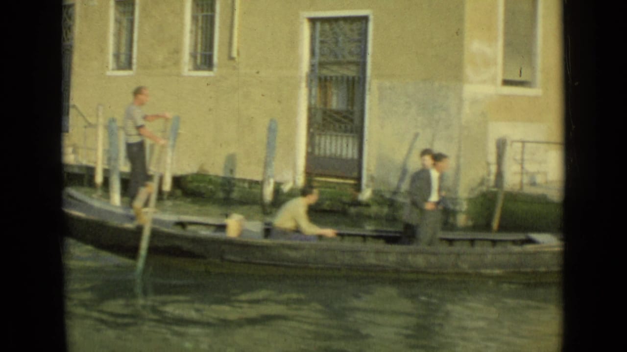 The gondolier stands with an oar while another person works low in the boat near a Venice canal landing.