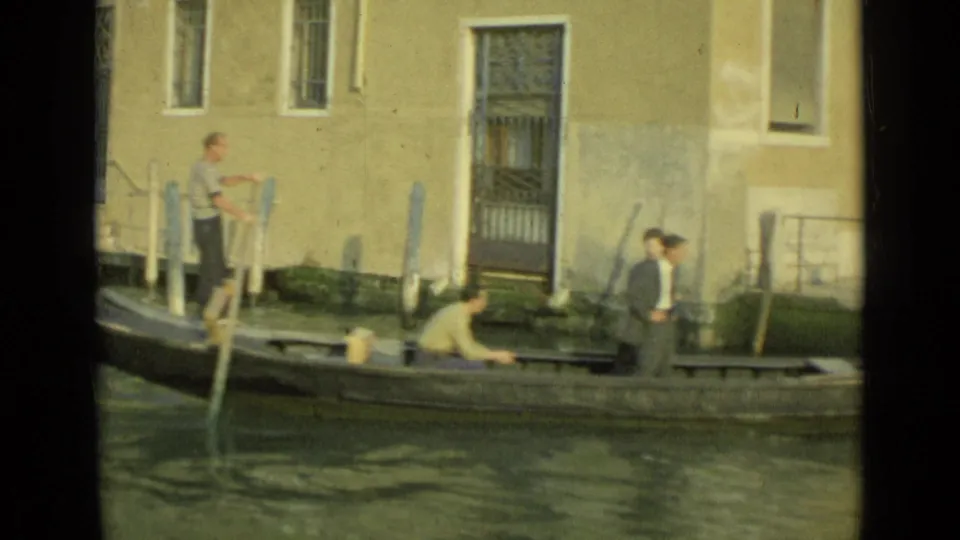 The gondolier stands with an oar while another person works low in the boat near a Venice canal landing.
