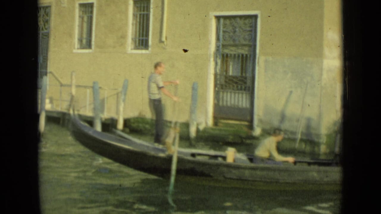 1962 Venice home-movie frame showing a gondola near a waterfront building, with a gondolier standing and a passenger seated low in the boat.