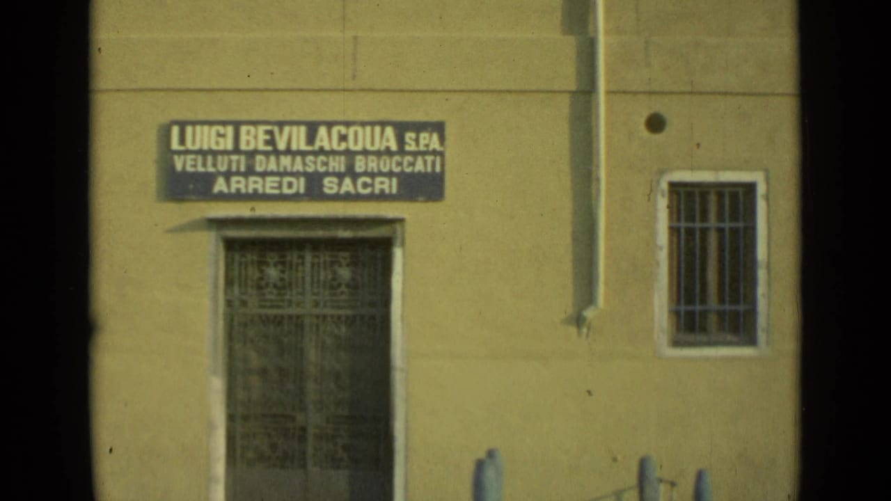 1962 Venice home-movie frame showing a waterfront facade with barred openings, blue mooring posts, and a visible Italian business sign.