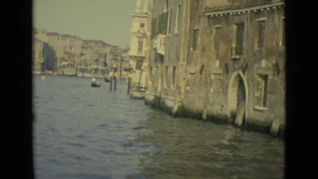 1962 Venice canal view looking along water between weathered buildings, with mooring poles and small boats in the distance.