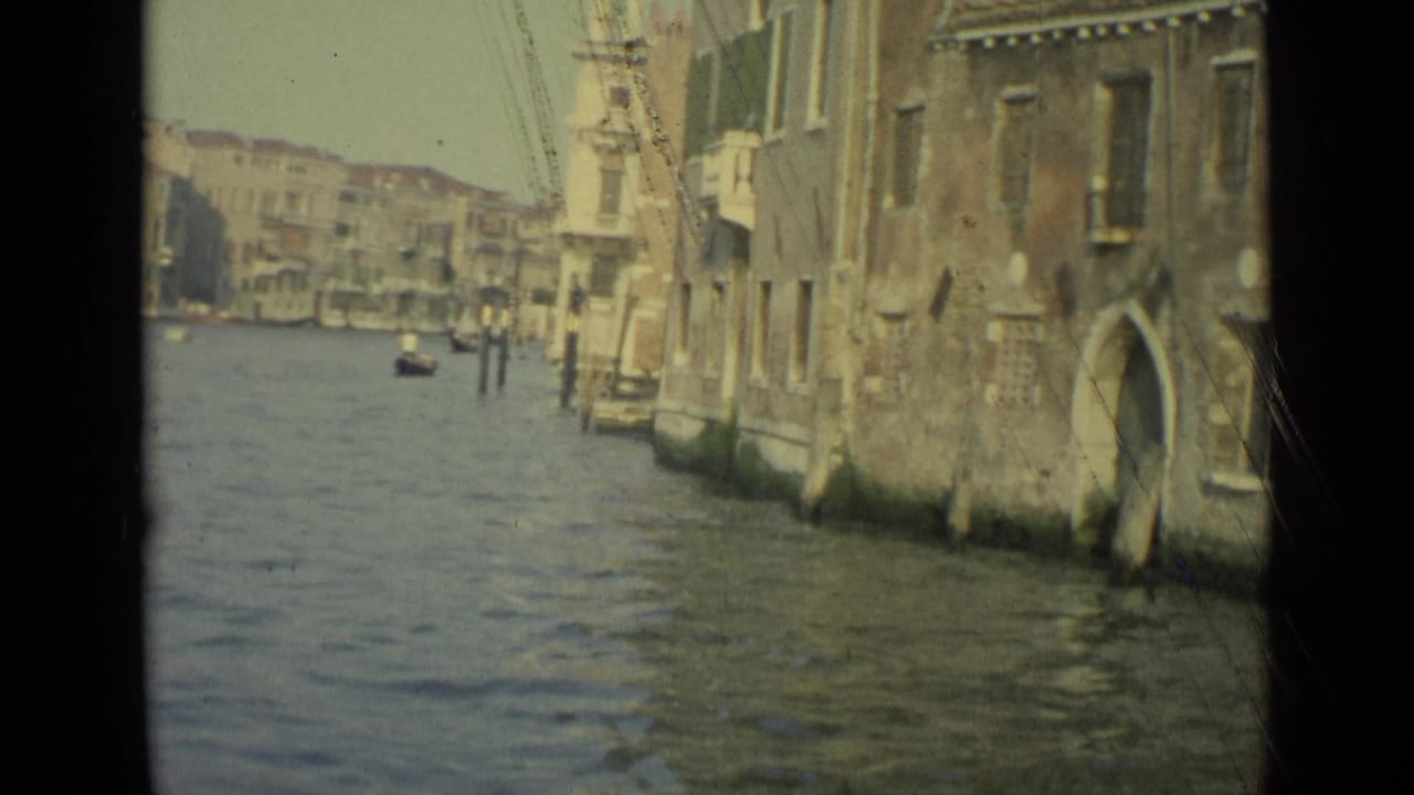 1962 Venice canal footage showing weathered waterfront buildings, arched openings, mooring posts, and rippling water from a boat-level view.