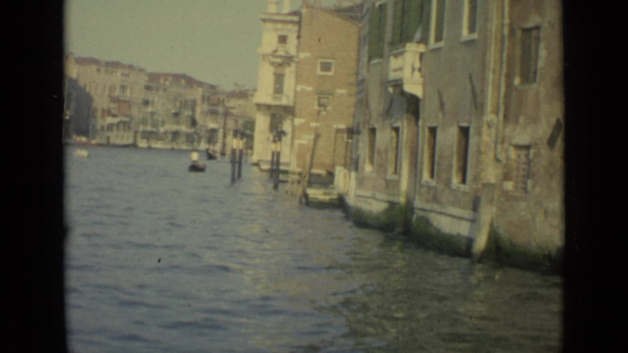1962 Venice home-movie frame showing canal water, weathered building walls, mooring posts, and distant waterfront structures.