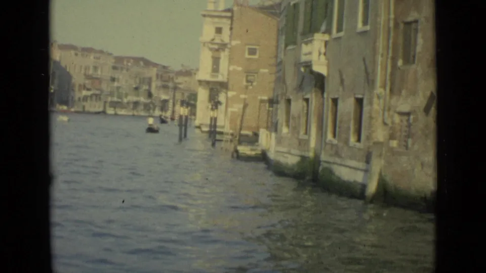 1962 Venice home-movie frame showing canal water, weathered building walls, mooring posts, and distant waterfront structures.